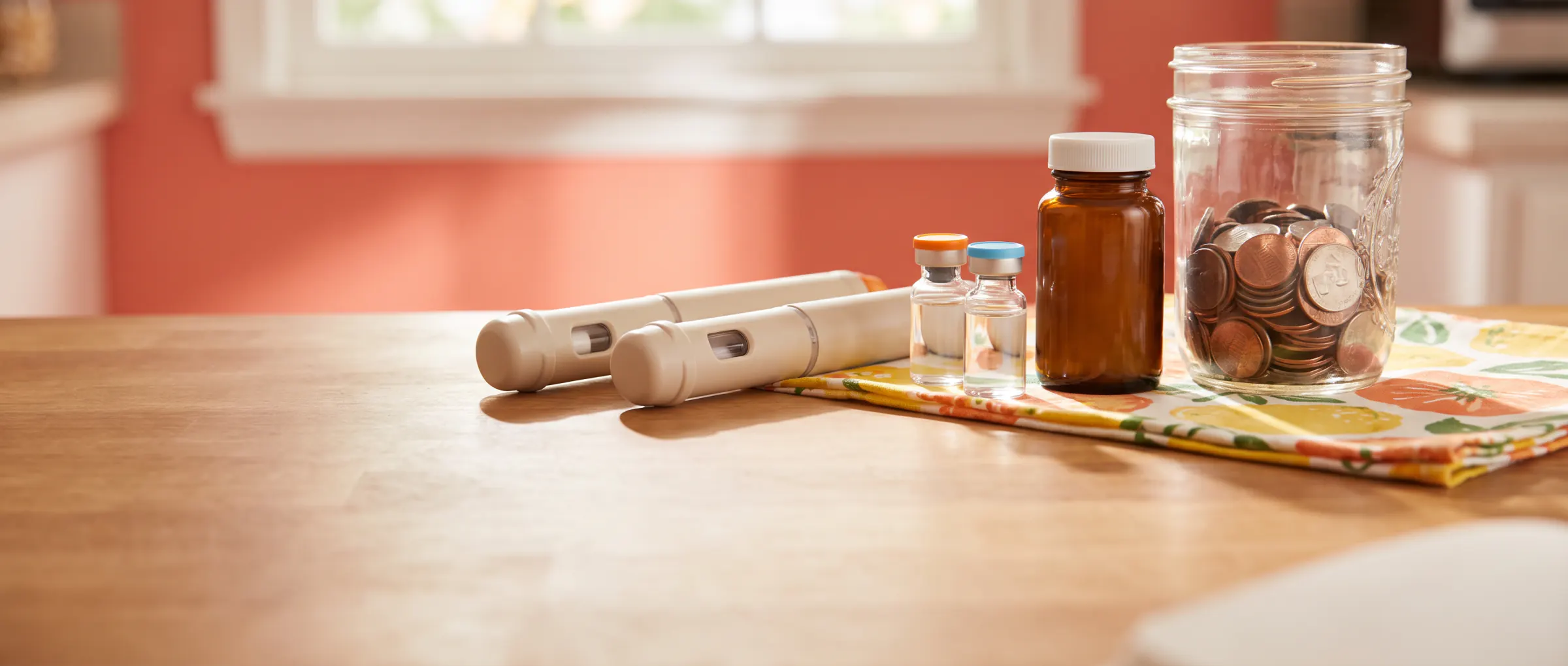 Plain injection pens, vials and a pill bottle beside a jar of coins on a sunny table