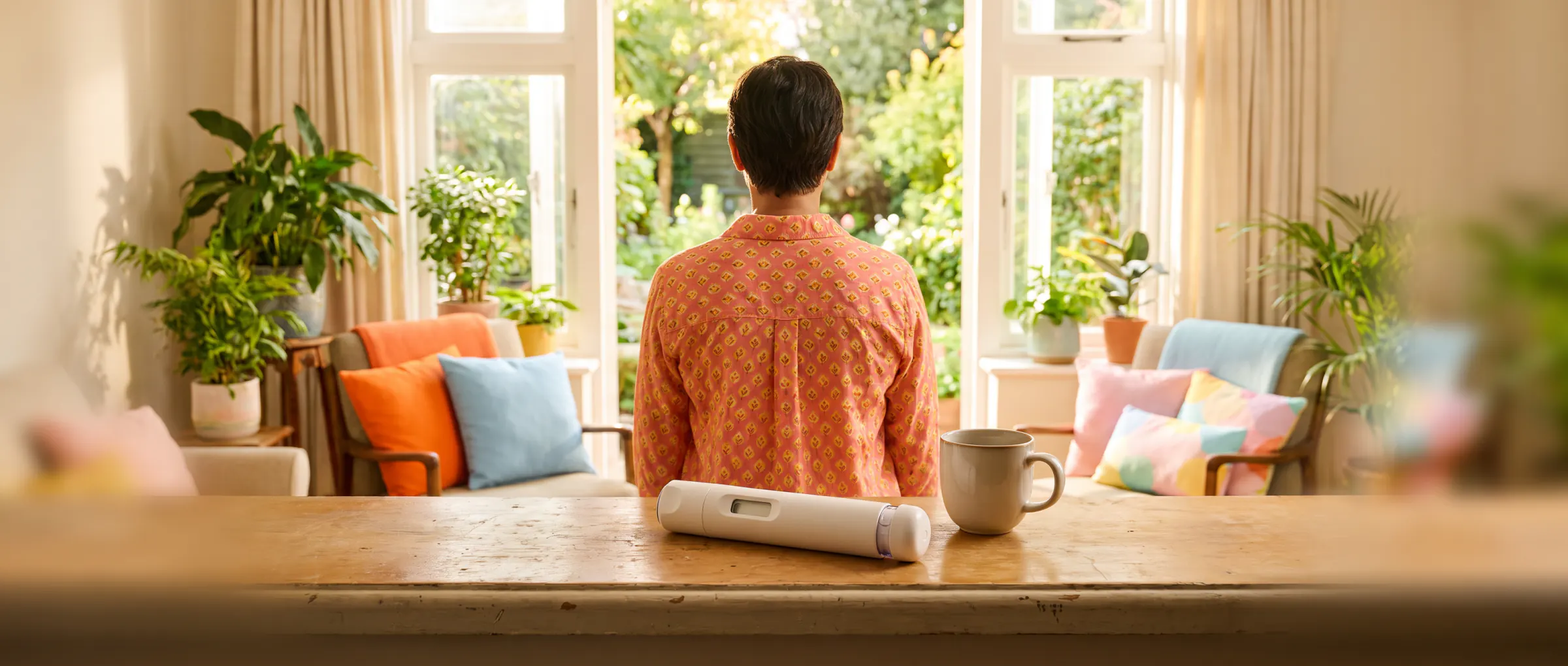 A person seen from behind at a sunny window with plants, a plain injection pen on the sill